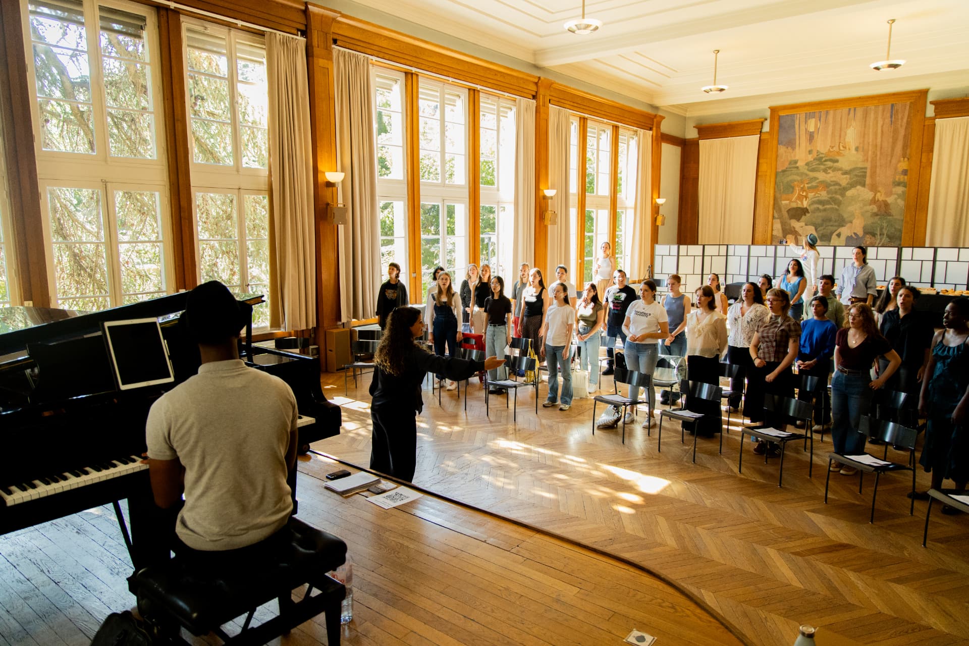 A pianist at a black grand piano in the foreground while the conductor directs standing singers in front of tall windows.