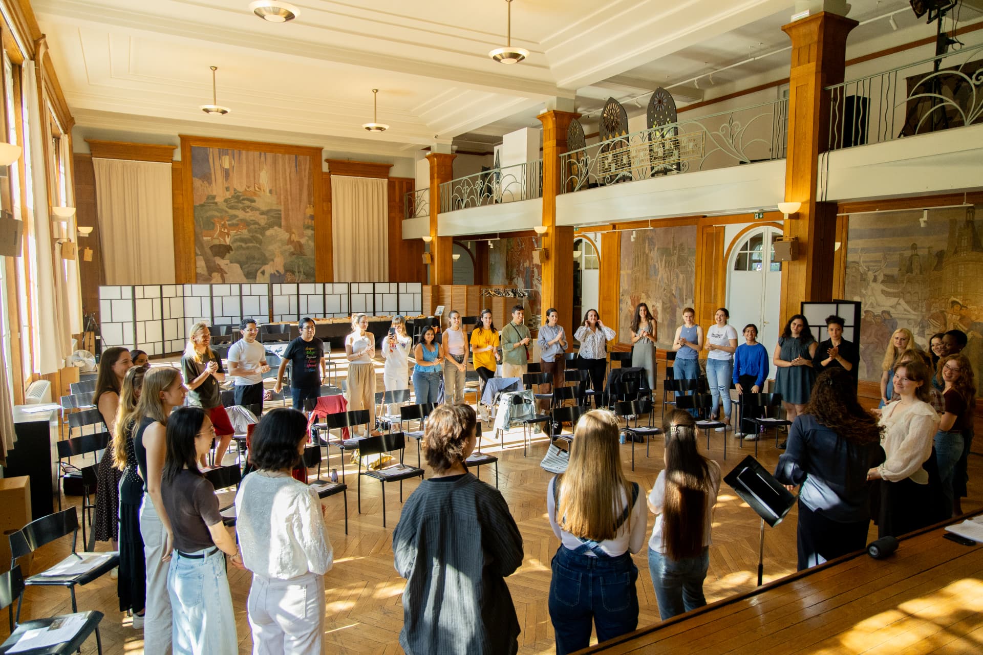 Singers stand in a circle around empty chairs in the Grand Salon, some clapping.