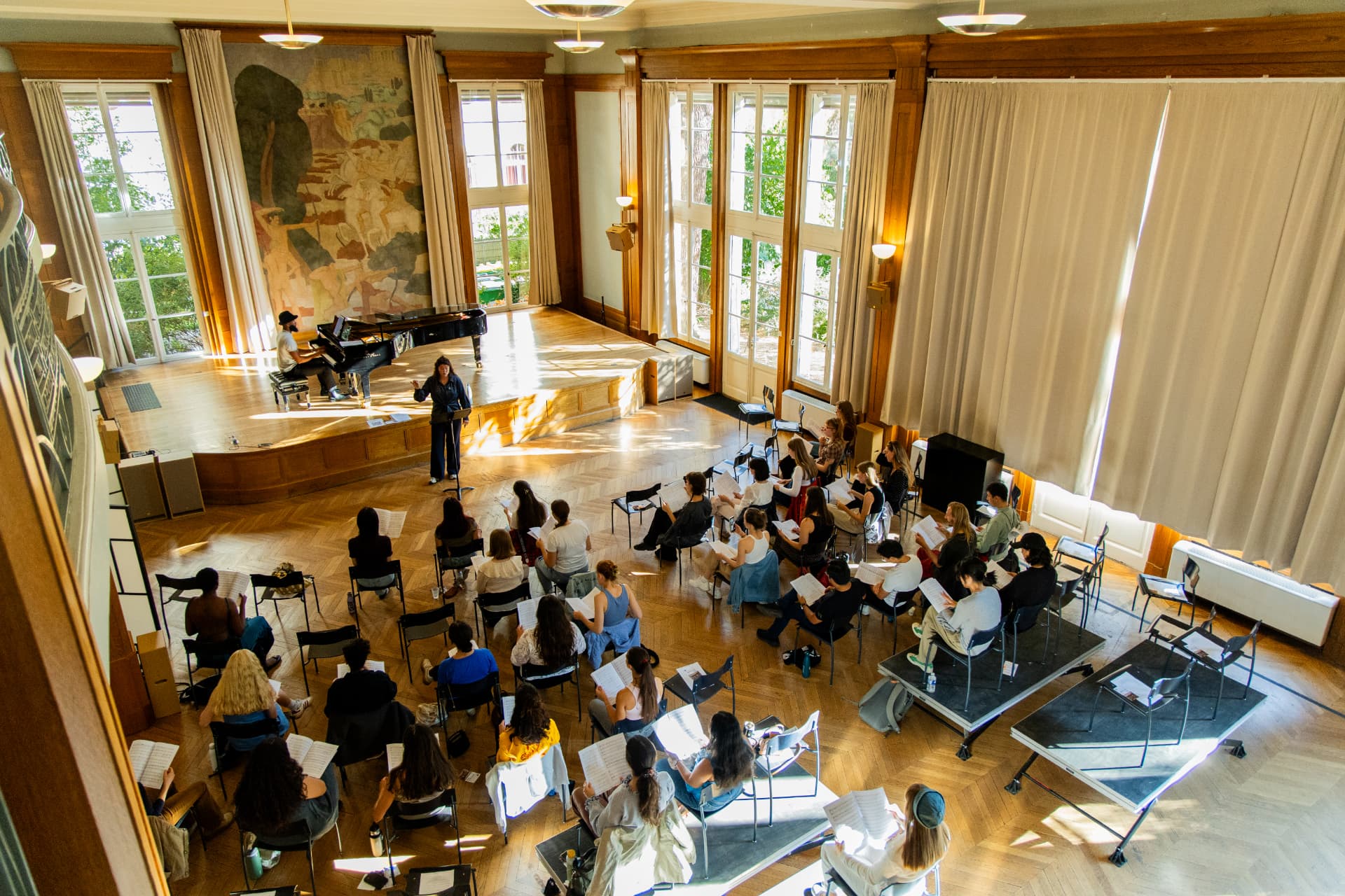 An elevated view of the Grand Salon: the conductor before rows of seated singers, a pianist at the grand piano beneath a wall mural.