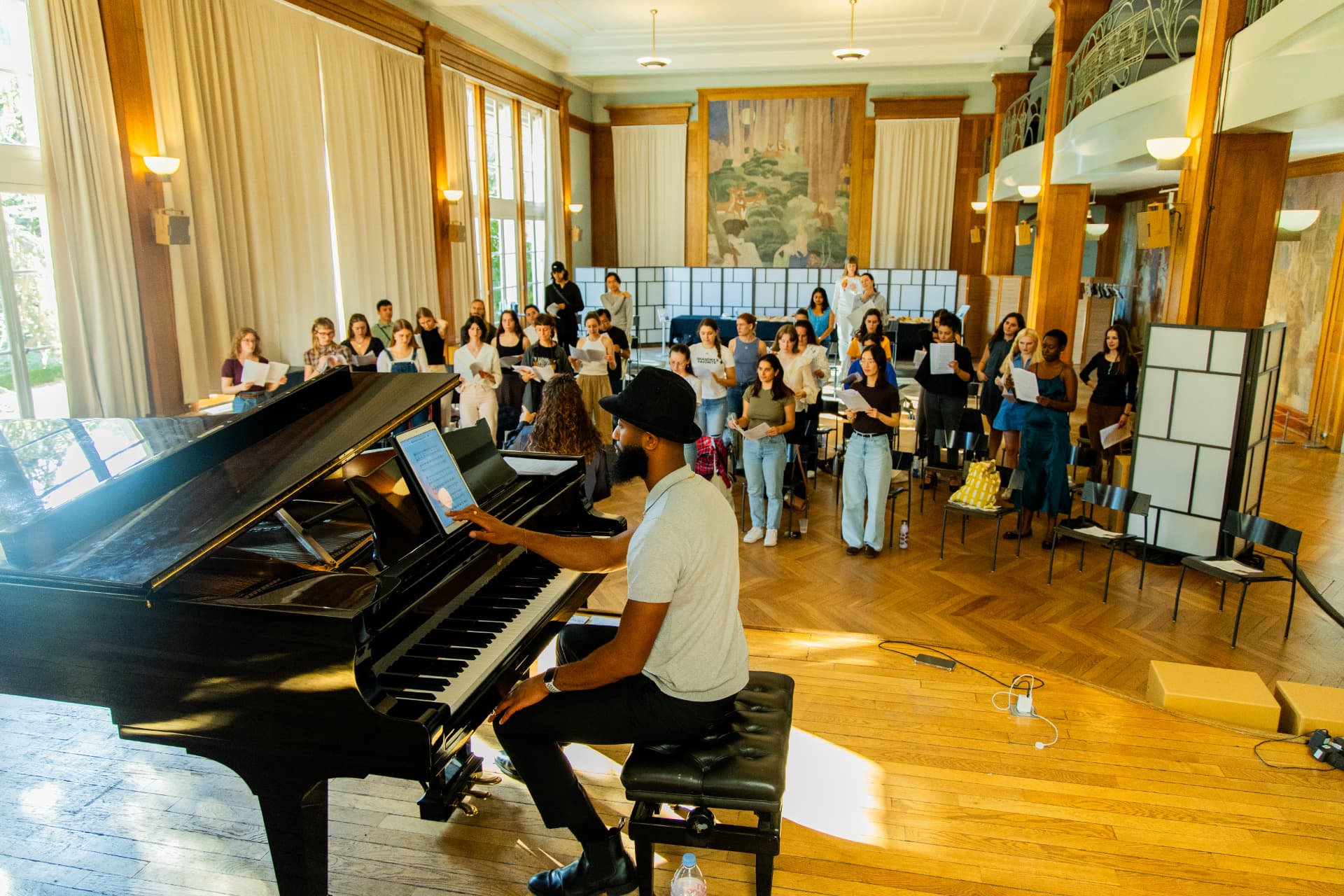 A pianist at the grand piano on the raised wooden stage faces standing singers holding sheet music.