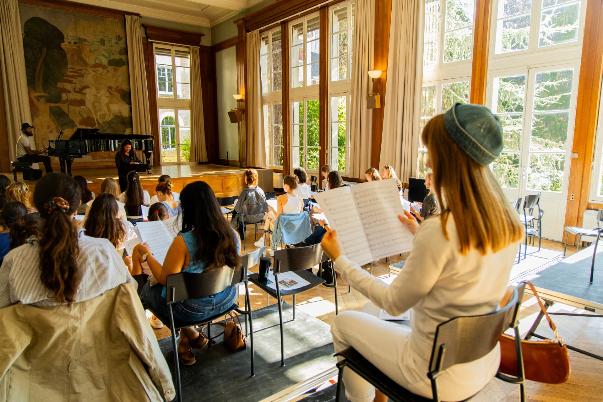 Singers seated with their backs to the camera hold open sheet music facing the conductor and pianist at the front of the hall.