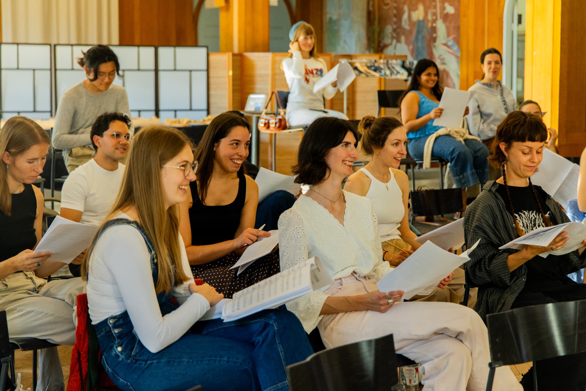 Four young singers sit close together holding sheet music on their laps, smiling during the rehearsal.