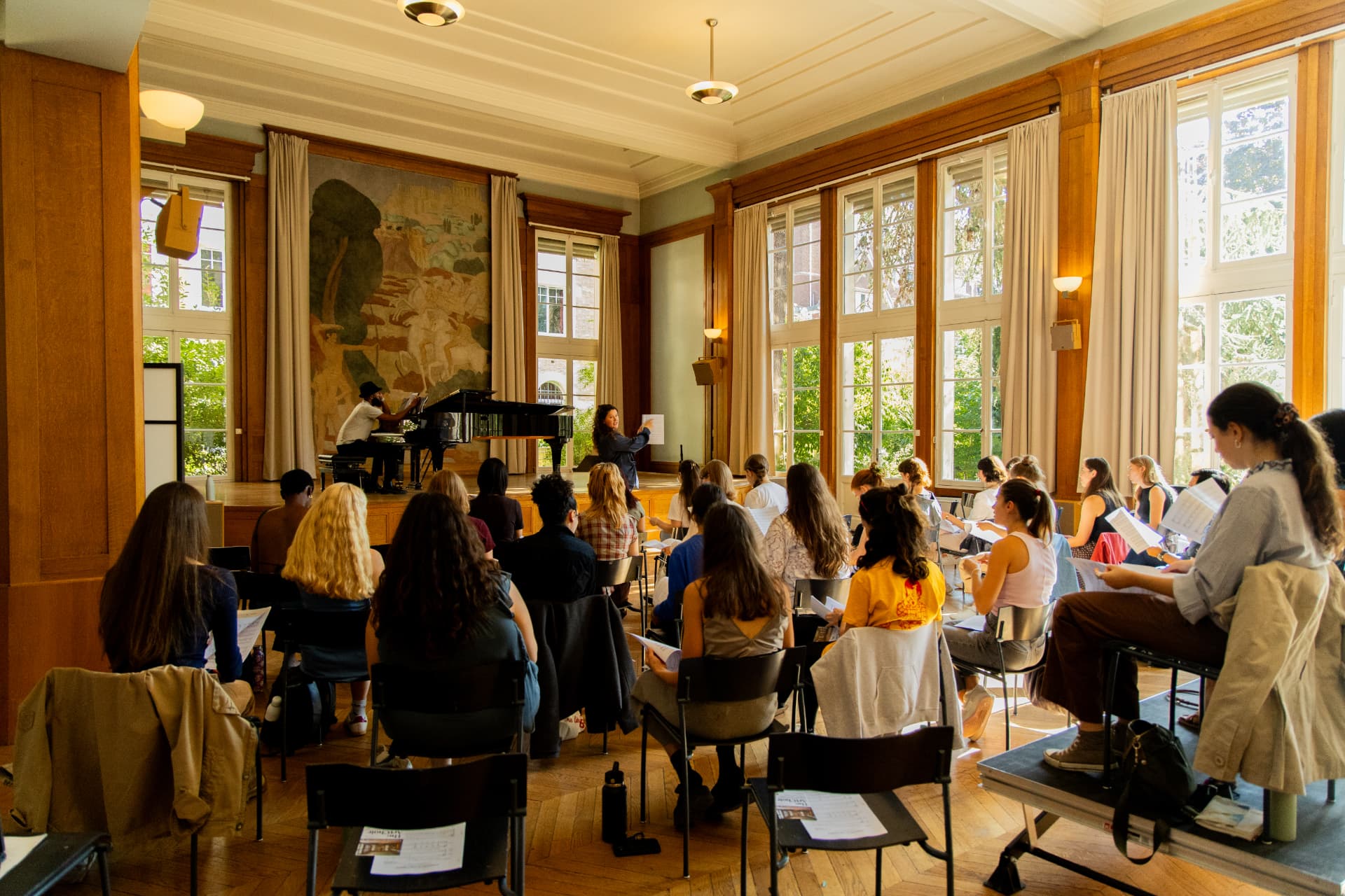 Singers seated in rows face the stage, where the conductor holds up sheet music beside a pianist at the grand piano.